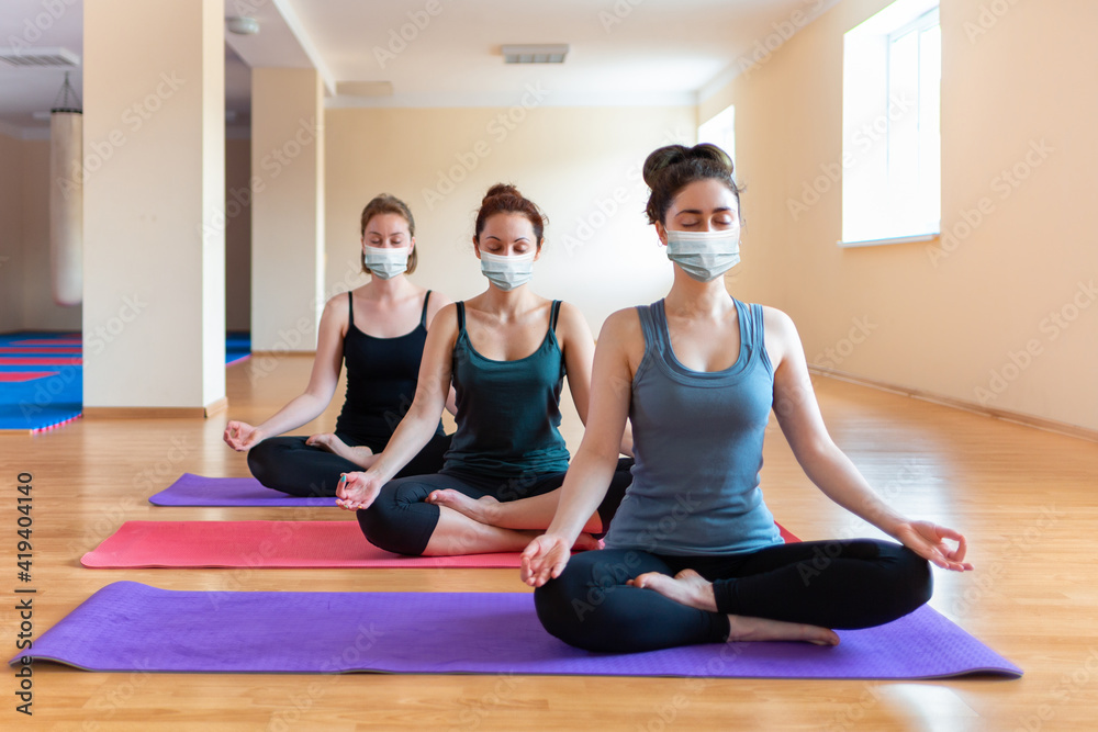 A group of young caucasian women in protective masks do yoga in the studio. The concept of a sporty lifestyle and new normal A group of young caucasian women in protective masks do yoga in the studio. The concept of a sporty lifestyle and new normal