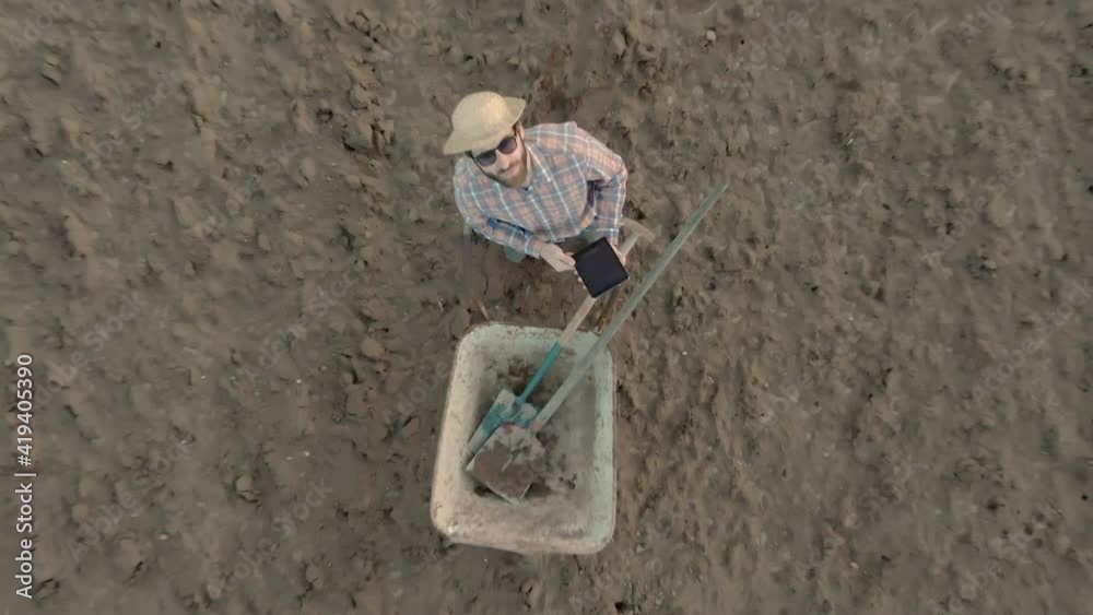 Aerial top view of male farmer working with tablet on farmland wearing straw hat plaid shirt rubber boots sunglasses at sunset shot in 4k Aerial top view of male farmer working with tablet on farmland wearing straw hat plaid shirt rubber boots sunglasses at sunset shot in 4k
