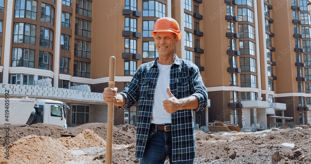 Fototapeta premium Smiling builder holding shovel and showing thumb up on construction site