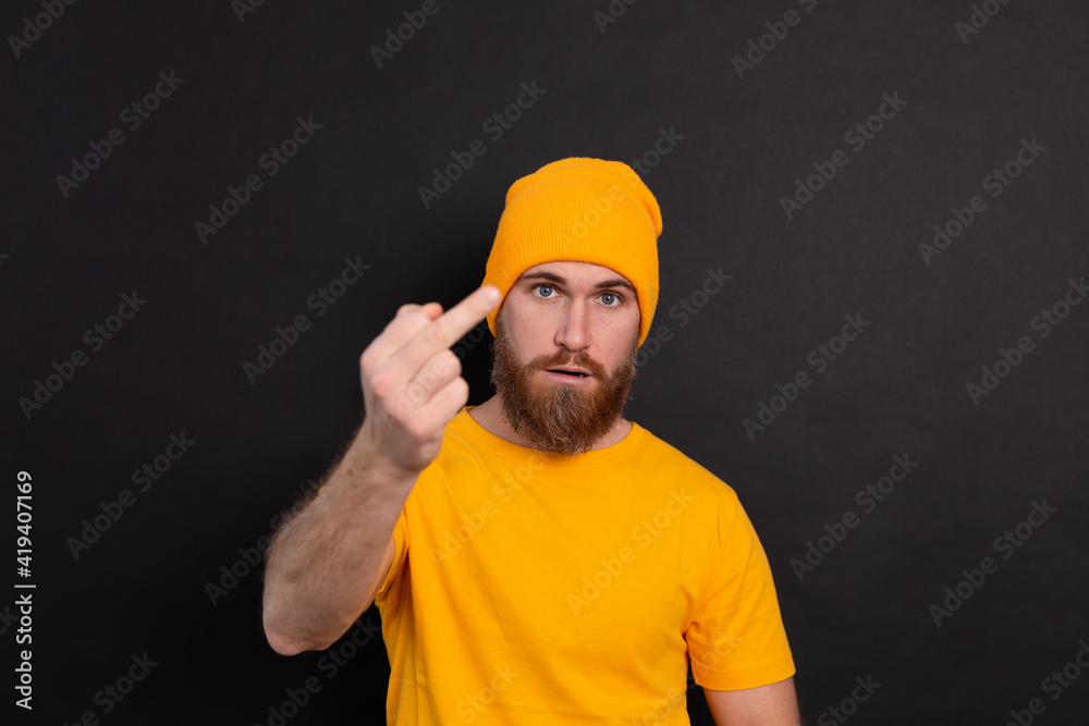 Portrait of bearded european man in yellow hat on black background serious look shows middle finger look to camera