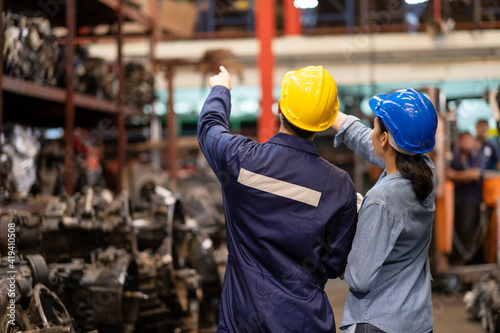 Two engineer or manager worker wearing safety helmet standing in the automotive part warehouse. Man and woman point forward to spare part in the rack. Inspector and quality check concept.