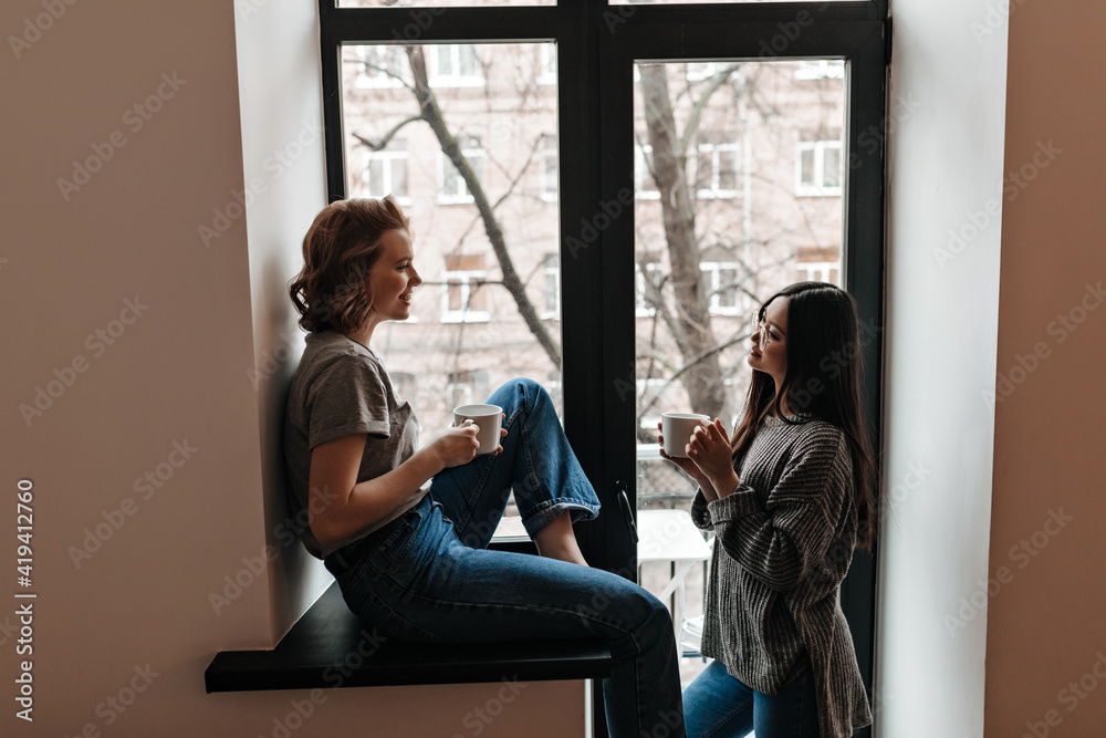 Two charming girls looking at each other while drinking coffee. Indoor shot of multiethnic friends enjoying tea near window. Two charming girls looking at each other while drinking coffee. Indoor shot of multiethnic friends enjoying tea near window.