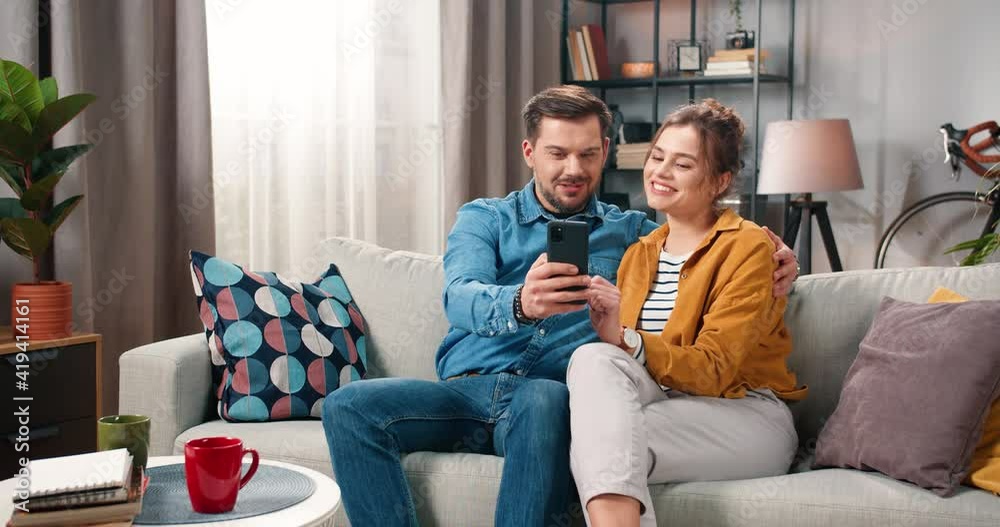 Lovely Caucasian young cute family couple wife and husband browsing online on smartphone resting at home sitting on sofa in modern living room, chatting and smiling spending time together love concept Lovely Caucasian young cute family couple wife and husband browsing online on smartphone resting at home sitting on sofa in modern living room, chatting and smiling spending time together love concept
