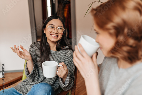 Selective focus of good-humoured friends drinking tea at home. Two charming girls talking and enjoying coffee.