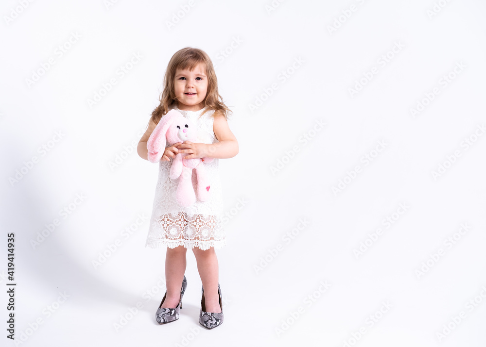 Funny little girl in white dress trying her mother shoes with heels on white background Funny little girl in white dress trying her mother shoes with heels on white background