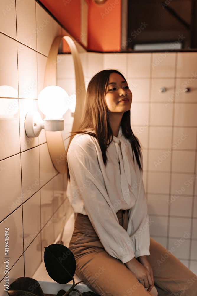 Smiling asian girl in white shirt looking away. Indoor shot of chinese pretty young woman in bathroom.