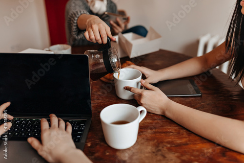 Cropped view of girls with laptop sitting at table during tea time. Partial shot of young woman pouring coffee to friends.