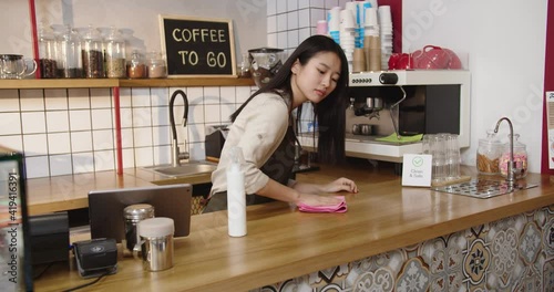 Hard-working female bartender wipping down bar in coffeehouse. Adult Asian woman waitrees disinfecting, sanitizing table surface, looking at camera and smiling in cafe. Virus, microbes concept. photo