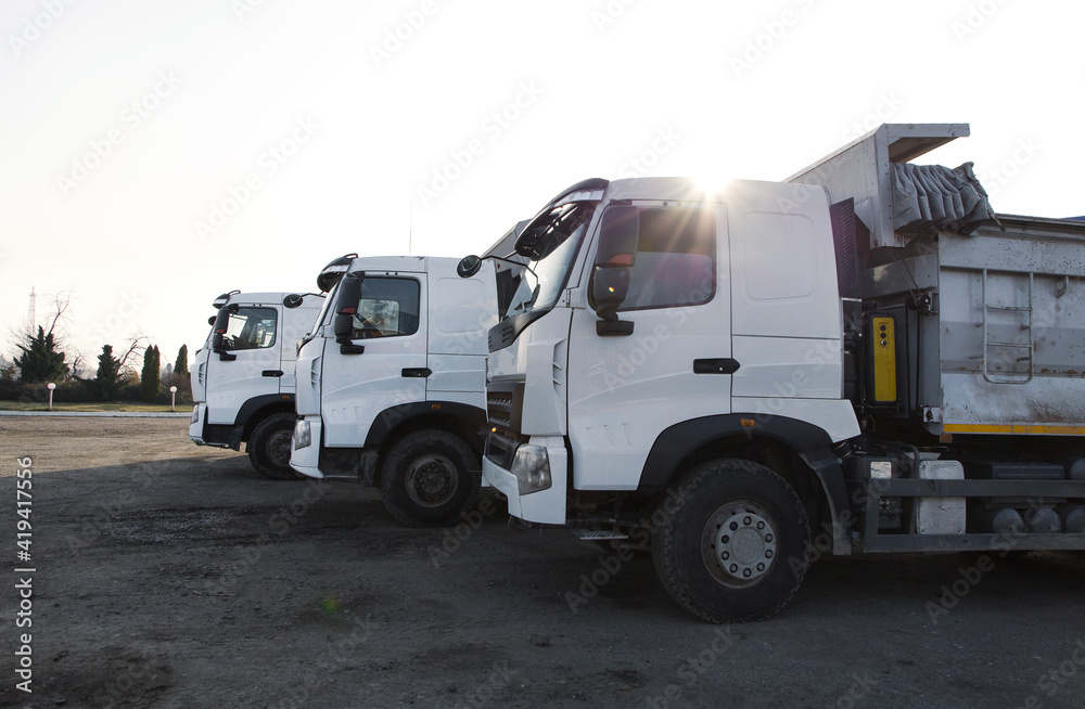 three dump trucks with white clean cabins stand next to each other. Delivery, logistics, construction, commercial transport. Sun rays. Copy space three dump trucks with white clean cabins stand next to each other. Delivery, logistics, construction, commercial transport. Sun rays. Copy space