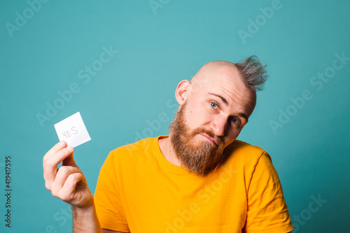 Bearded european man in yellow shirt isolated on turquoise background holding yes with surprise and amazed expression excited face.