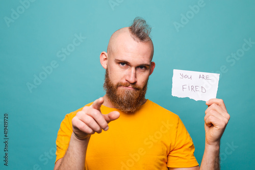 Bearded european man in yellow shirt isolated on turquoise background holding paper with you are fired text pointing with finger to the camera and to you, hand sign