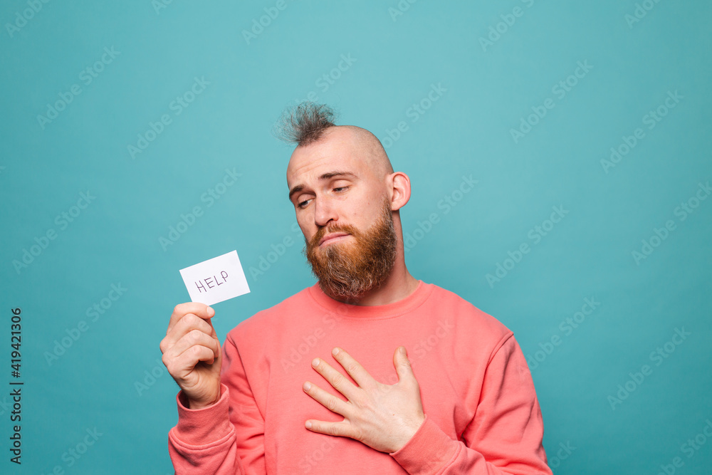 Bearded european man in casual peach isolated on turquoise background holding paper with help sign sad unhappy begs Bearded european man in casual peach isolated on turquoise background holding paper with help sign sad unhappy begs