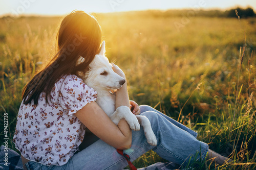 Photography Woman cuddling with cute white puppy in summer meadow and looking at warm sunset light