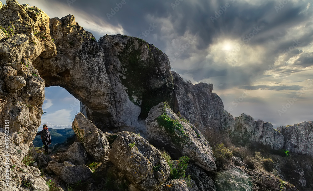 Panoramic view of a big arch in the summit of a hill. Beams of sun coming through the clouds. One girl is standing inside the arch