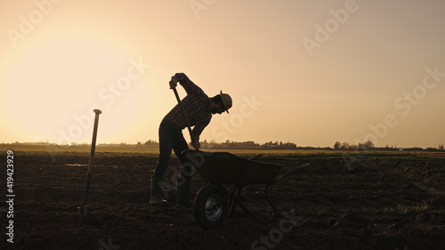 Male farmer shovels earth in wheelbarrow on farmland wearing straw hat plaid shirt rubber boots sunglasses at sunset