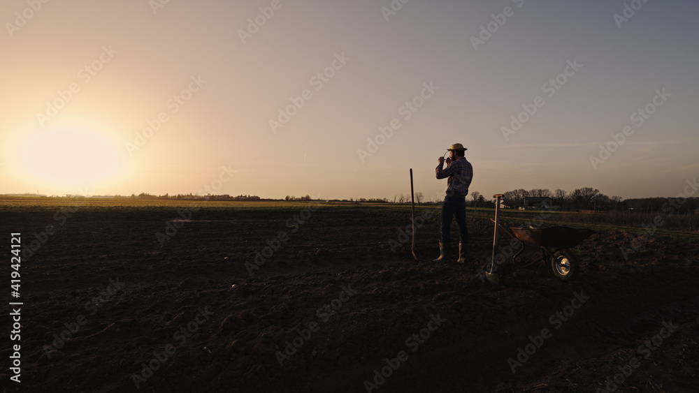 Male farmer making a rest of pushing wheelbarrow over farmland wearing straw hat plaid shirt rubber boots sunglasses at sunset