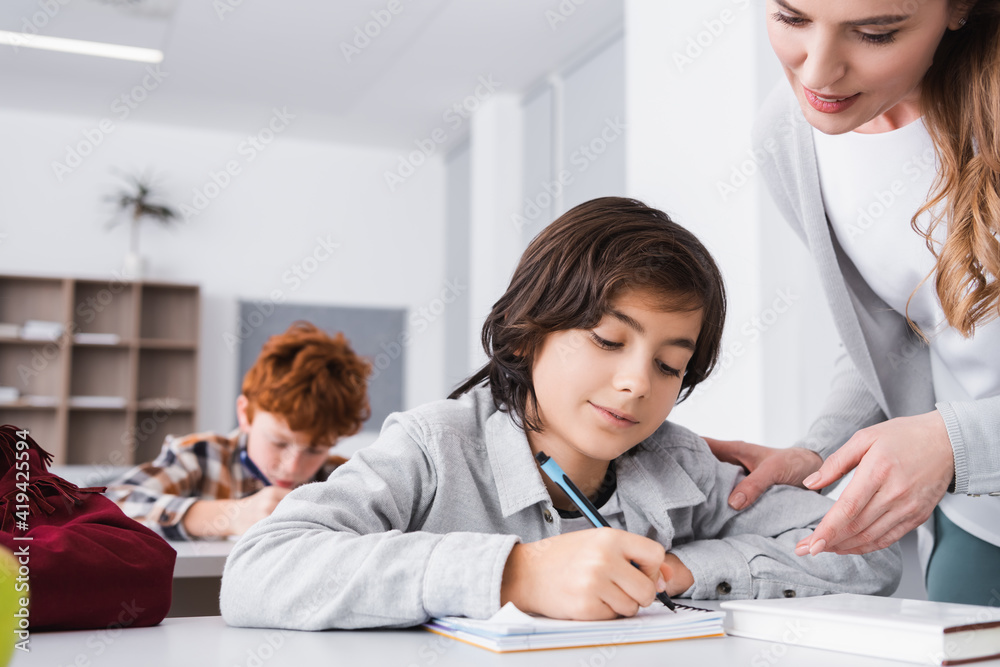 schoolboy writing in notebook near teacher helping him during lesson schoolboy writing in notebook near teacher helping him during lesson