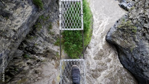 Pov: Person Steps Gingerly Across Narrow Rope Bridge Over Roaring River And Sharp Cliffs photo