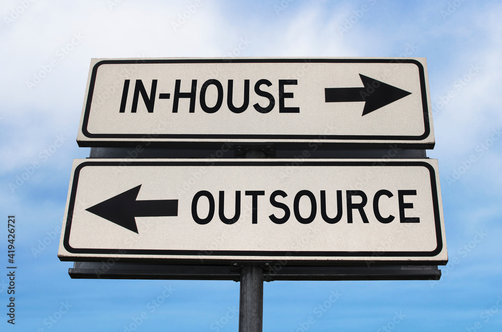 Outsource versus in-house road sign with two arrows on blue sky background. White two streets sign with arrows on metal pole. Directional sign. Outsource versus in-house road sign with two arrows on blue sky background. White two streets sign with arrows on metal pole. Directional sign.