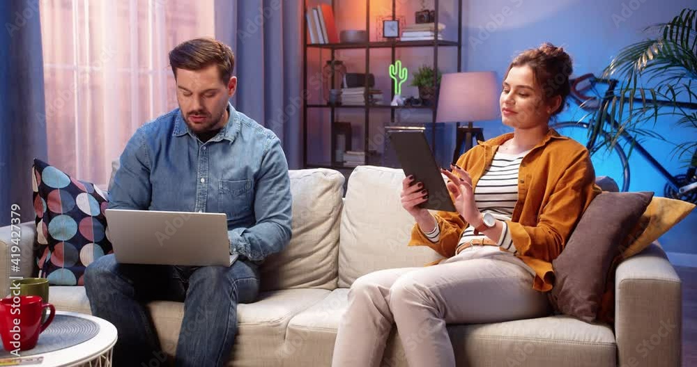 Caucasian young married couple sitting on couch in living room in evening spending time at home speaking and using digital gadgets Man browsing online on laptop while his pretty wife tapping on tablet