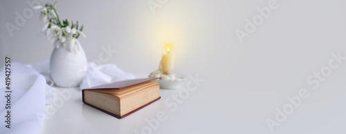 panorama, banner of still life in high key style, old paper book in a dark red binding on white table, candle, bouquet of half snowdrops in vase, concept of changing seasons, reading literature