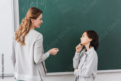 thoughtful schoolboy looking at smiling teacher near chalkboard