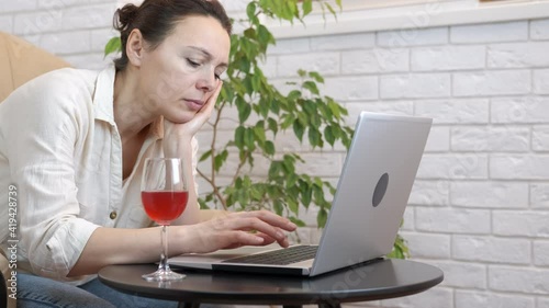 Freelancer with drinking problem. A woman with a glass is sadly working on a laptop at home. At home office with a glass of wine. photo