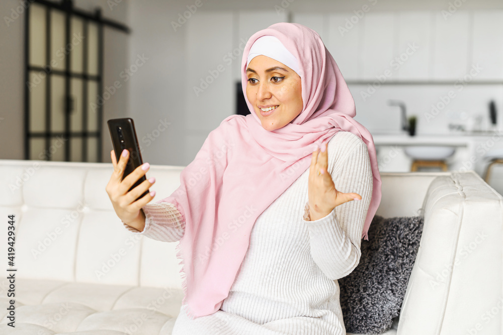 Young Muslim Arabic mixed-race woman talking online on mobile phone, sitting on the couch in studio apartment, video-calling with family coworkers, chatting, gesturing, discussing news with a friend