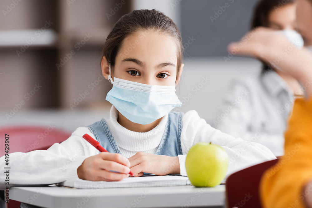 schoolgirl in medical mask looking at camera near apple and classmate on blurred foreground