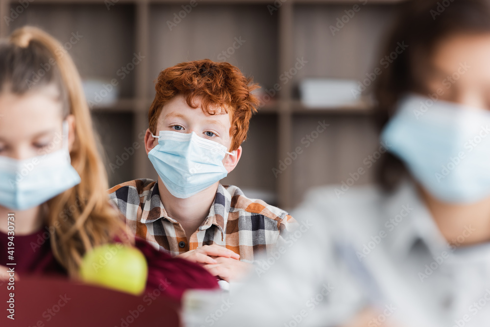 redhead boy in medical mask looking at camera during lesson, blurred foreground