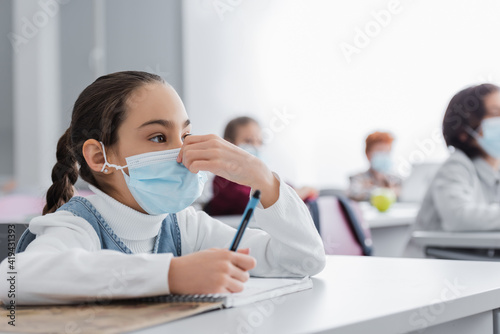 schoolgirl touching medical mask while holding pen during lesson