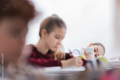 Schoolchild using smartphone near classmates on blurred foreground in class