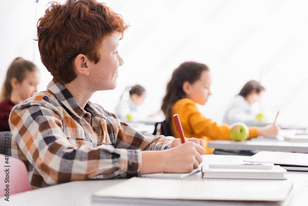 Schoolboy writing on notebook near laptop on blurred foreground in classroom Schoolboy writing on notebook near laptop on blurred foreground in classroom