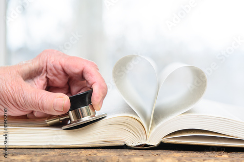 An open book. The pages are folded in the shape of a heart. In the hands of an elderly woman, a stethoscope is pressed against the pages. She measures the pulse of the book's heart. Concept-education,