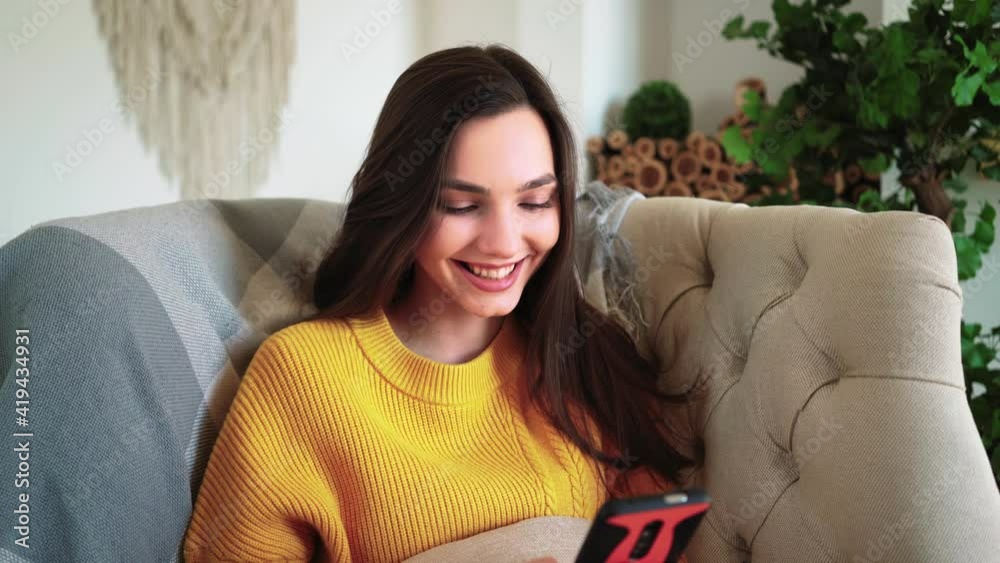 Relaxed Woman in Yellow Sweater Smiling Hold Smartphone Watching Social Media Stories Video Sit on Sofa Home. Happy Girl Look Cellphone Laughing Enjoying Using Mobile App Having Fun Play and Chatting Relaxed Woman in Yellow Sweater Smiling Hold Smartphone Watching Social Media Stories Video Sit on Sofa Home. Happy Girl Look Cellphone Laughing Enjoying Using Mobile App Having Fun Play and Chatting