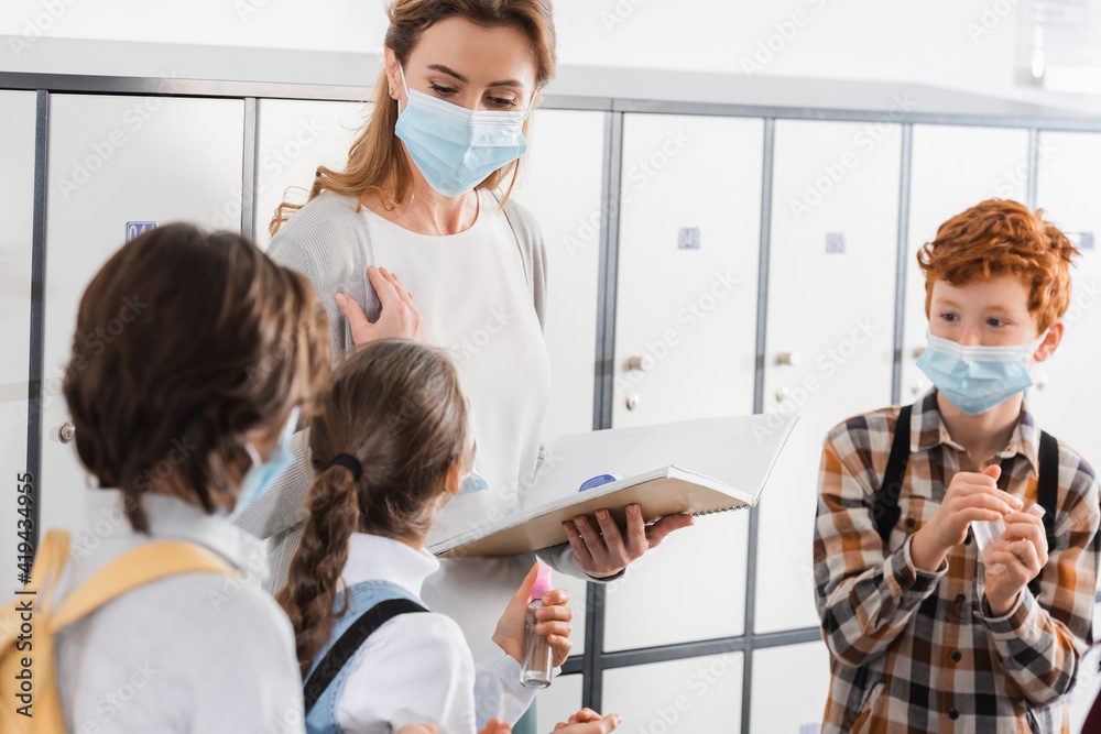 Pupils holding hand sanitizer near teacher in medical mask with notebook and infrared thermometer Pupils holding hand sanitizer near teacher in medical mask with notebook and infrared thermometer