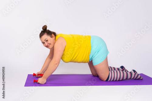 Caucasian plump woman on a fitness mat with dumbbells trying to lose weight. Exercises for the body. Sports at home in comfortable clothing on a gray background.