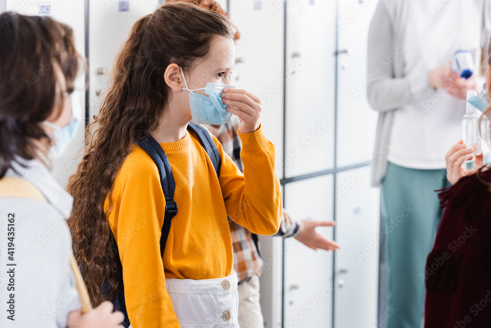 Schoolgirl wearing medical mask near friends with hand sanitizer on blurred foreground Schoolgirl wearing medical mask near friends with hand sanitizer on blurred foreground