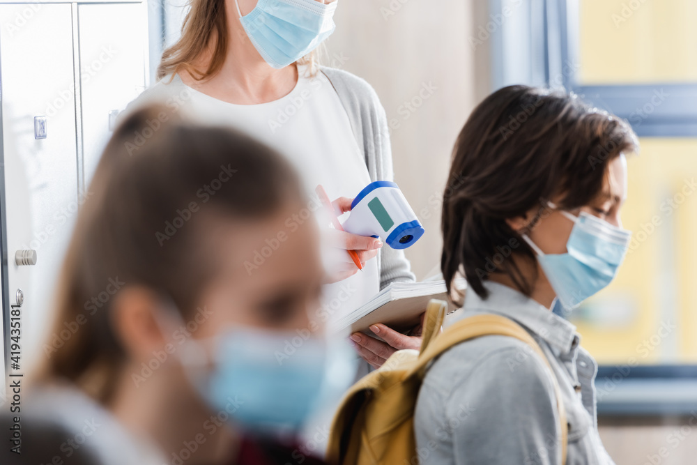 Teacher holding infrared thermometer and notebook near pupils in medical masks on blurred foreground Teacher holding infrared thermometer and notebook near pupils in medical masks on blurred foreground