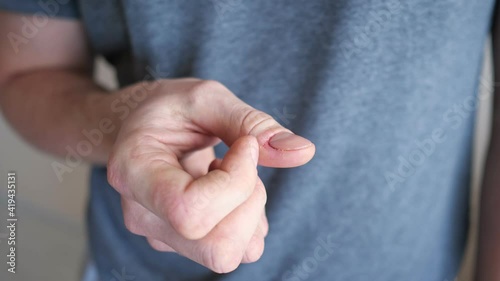 Man in grey t-shirt scratches agnail on finger suffering from neurosis on blurred beige background in light room extreme close crop view