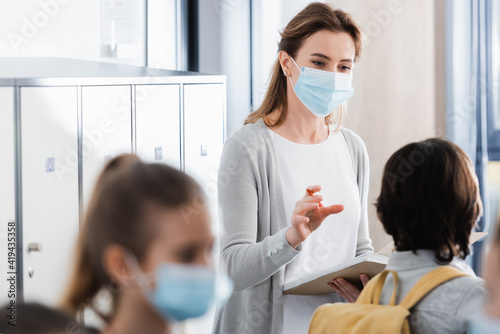 Teacher in medical mask holding notebook and looking at pupil on blurred foreground