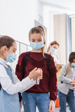 Schoolkid in protective mask looking at friend with hand sanitizer on blurred foreground