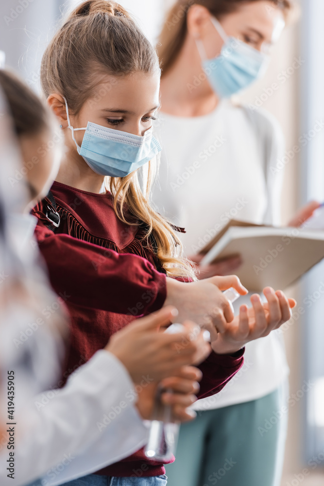 Pupil in protective mask using hand sanitizer near friend and teacher on blurred background