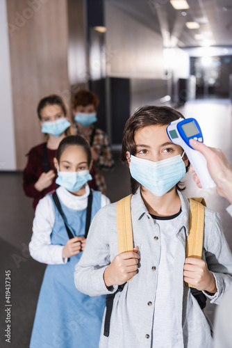 Teacher with non contact thermometer measuring temperature of schoolboy near kids in medical masks in school