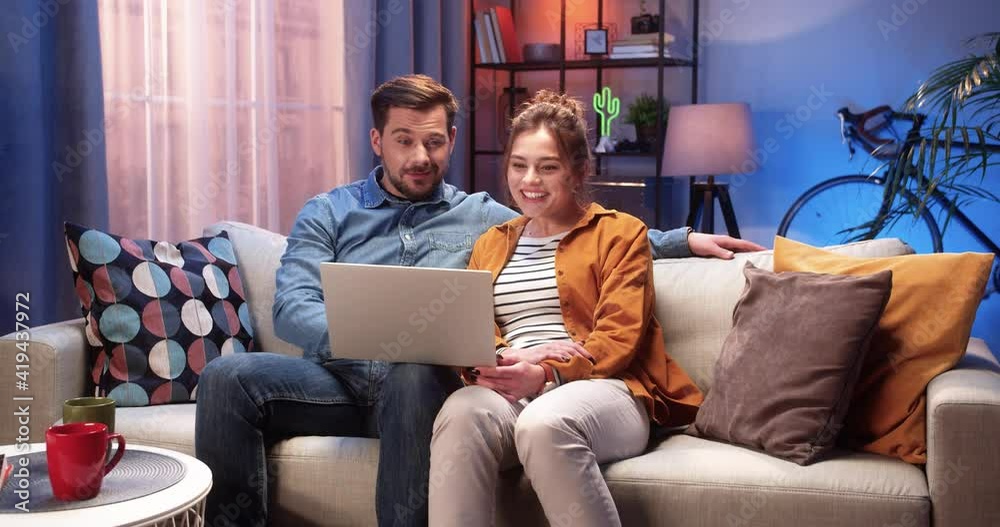 Young Caucasian lovely couple handsome husband speaking with beautiful wife sitting on couch in cozy room in house searching internet on laptop computer choosing something Family time leisure concept Young Caucasian lovely couple handsome husband speaking with beautiful wife sitting on couch in cozy room in house searching internet on laptop computer choosing something Family time leisure concept