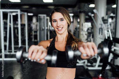 Beautiful sports girl in the gym reaches out with dumbbells to the camera, she smiles and looks at the camera, lifestyle