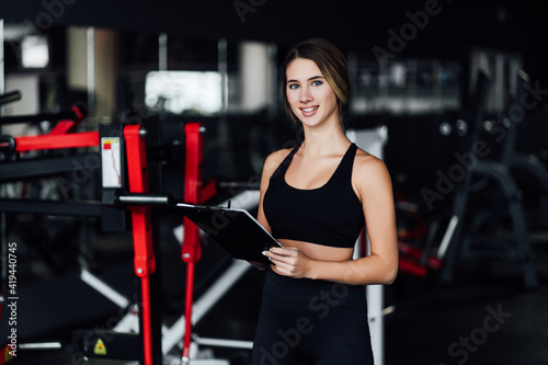 Attractive sports girl, personal trainer in the middle of a modern gym with a workout plan in her hands, she smiles and looks at the camera