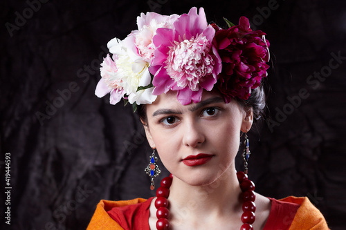 The beautiful girl brunette with red lips with a wreath of peonies on his head in the ethnic image of Mexican artist Frida Kahlo at dark background