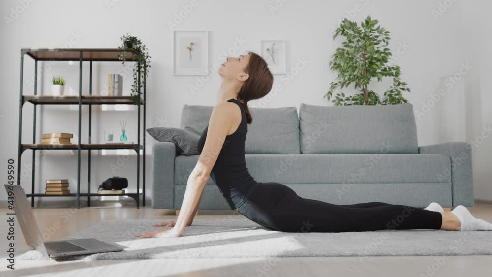 Side view of happy active woman stretching body on floor while watching online tutorial on modern laptop. Pretty brunette in sport clothes doing fitness exercises at home. Side view of happy active woman stretching body on floor while watching online tutorial on modern laptop. Pretty brunette in sport clothes doing fitness exercises at home.