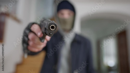 Close-up of gun muzzle with blurred masked man at background aiming. Unrecognizable Caucasian burglar thief threatening wealthy house owner with weapon. Stealing and robbing concept.